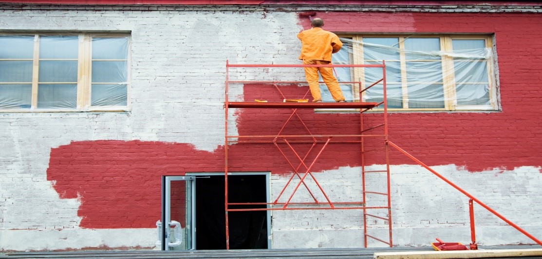 worker in an orange jumpsuit is painting the upper part of a wall red